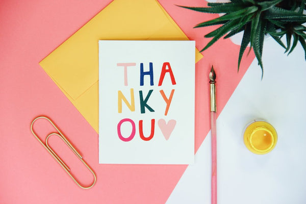 A white thank you card with the words "thank you" written in different colors sitting on top of a yellow envelope next to a paper clip, a plan, and a quilt pen